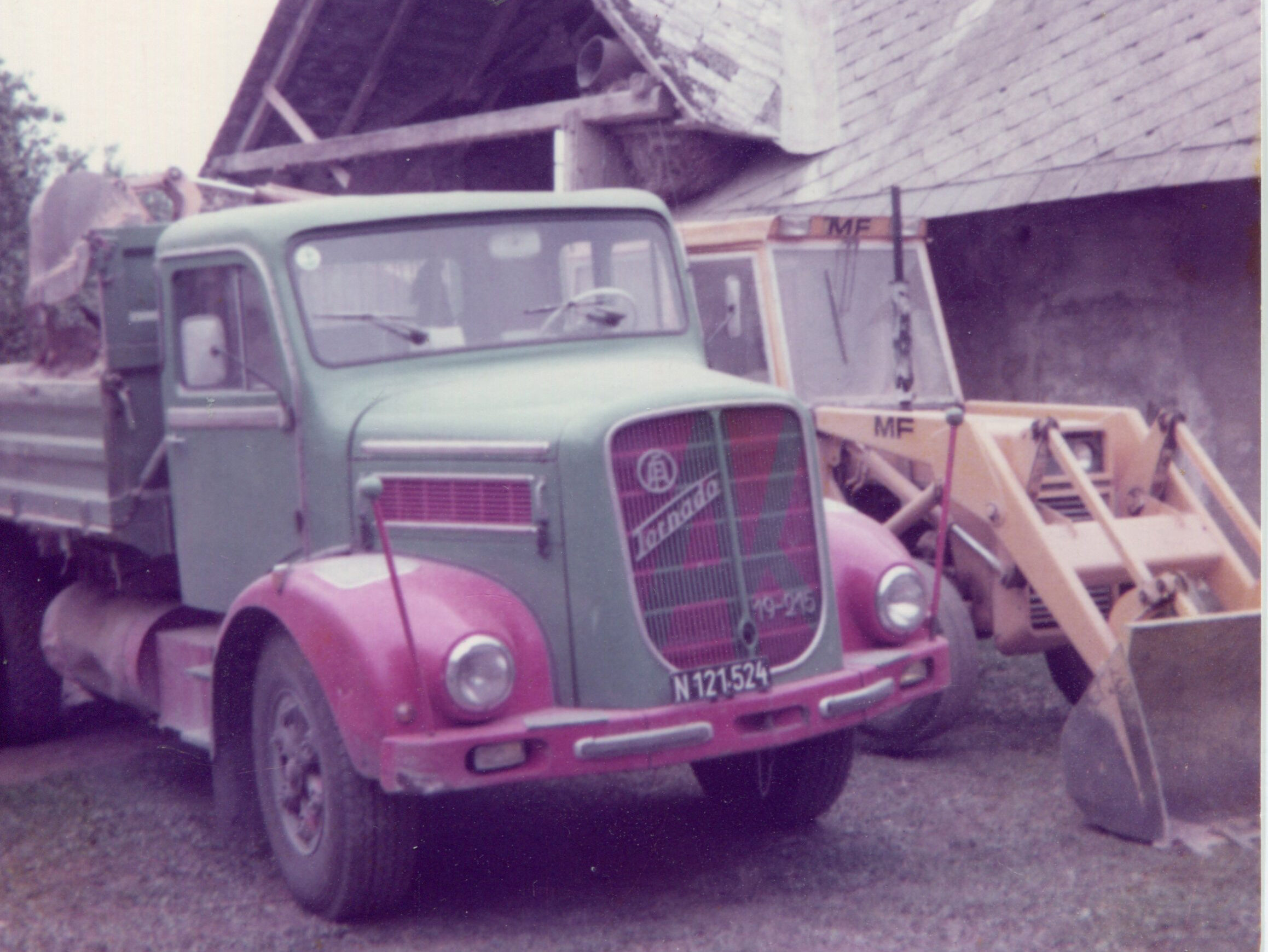 Grün-roter Oldtimer Lkw neben einem gelben Bagger vor Haus. 
