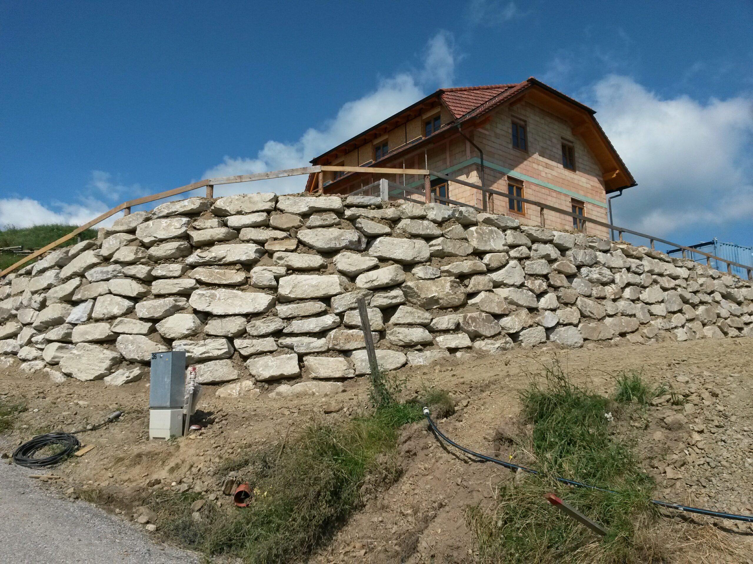 Massive Trockenmauer aus großen Natursteinen als Stützmauer am Hang; darüber ein Haus im Rohbau mit rotem Ziegeldach, blauer Himmel und Wolken.