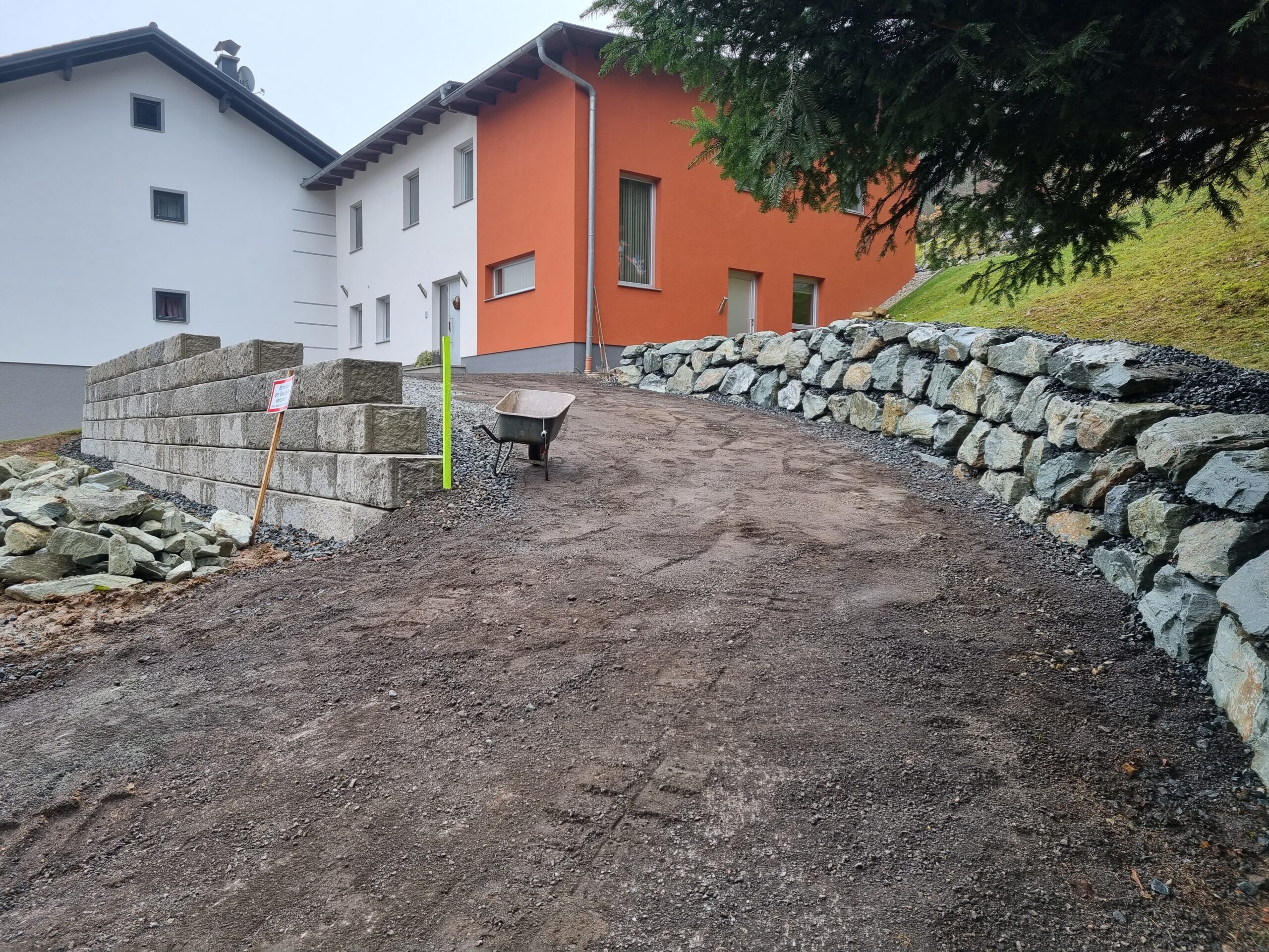 Eine Natursteinmauer neben einer Zufahrt zu einem Haus mit rot-weißer Fassade. 