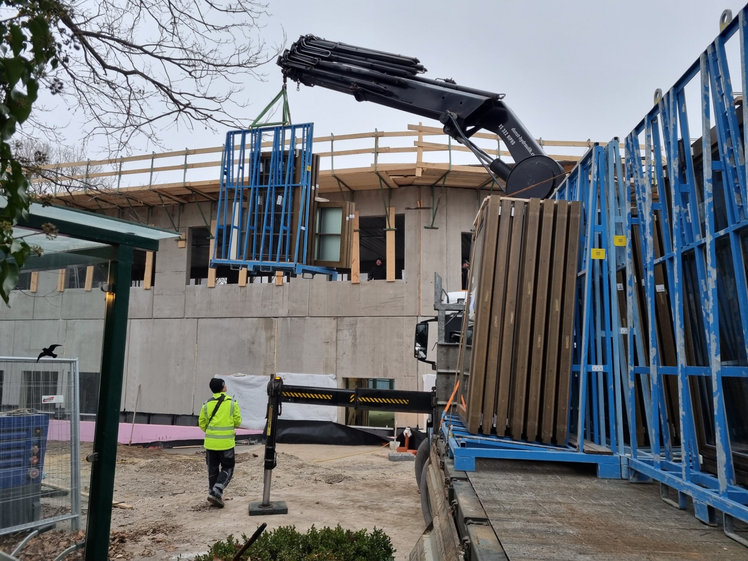 Schwarzer Ladekran hebt ein blaues Transportgestell mit Fenstern vor einem mehrstöckigen Betonrohbau; unten steht ein Arbeiter in Warnjacke, rechts ein Lkw mit weiteren Gestellen.