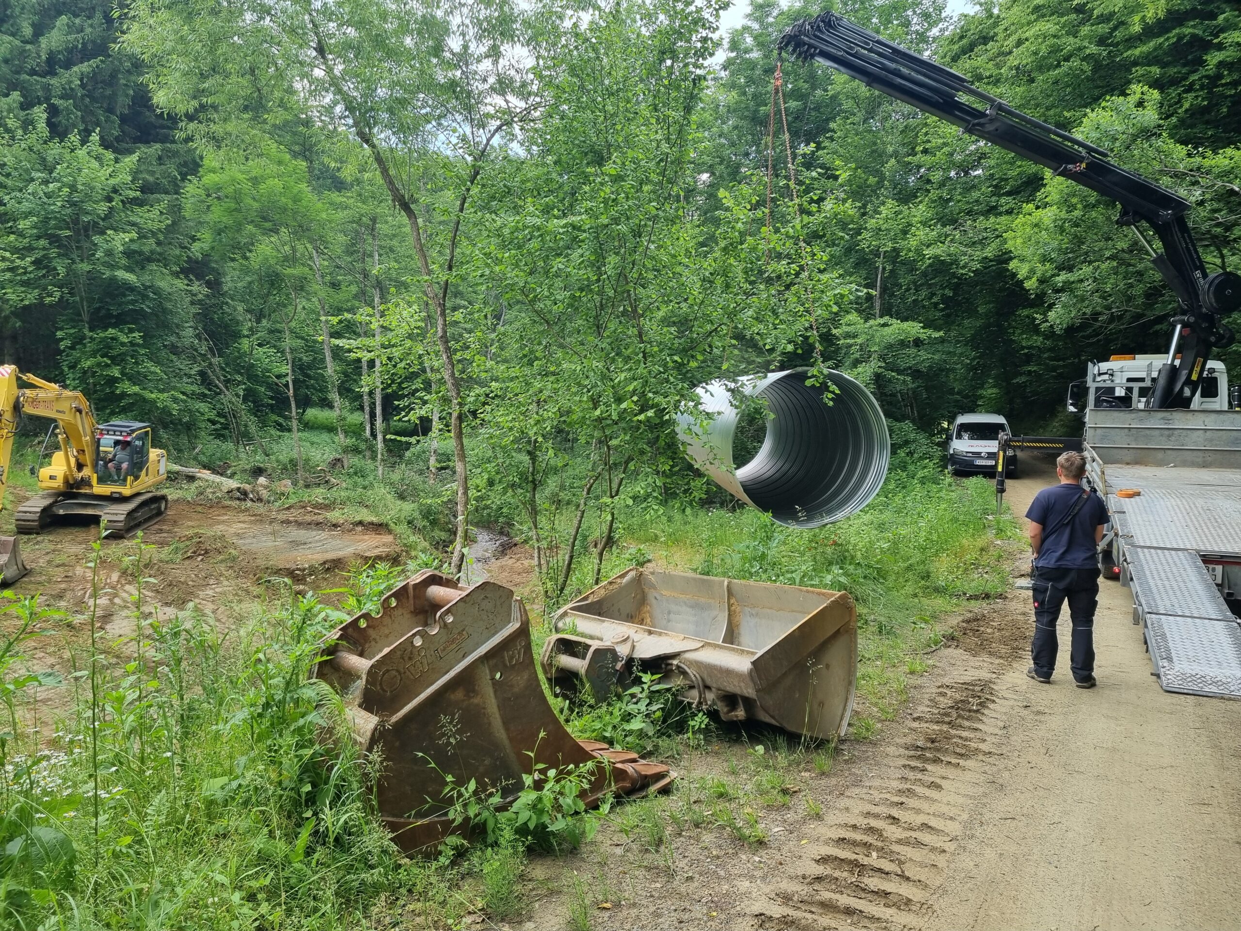 Ein Bagger saniert ein Bachbett neben einem Kran-Lkw auf einer Waldstraße.