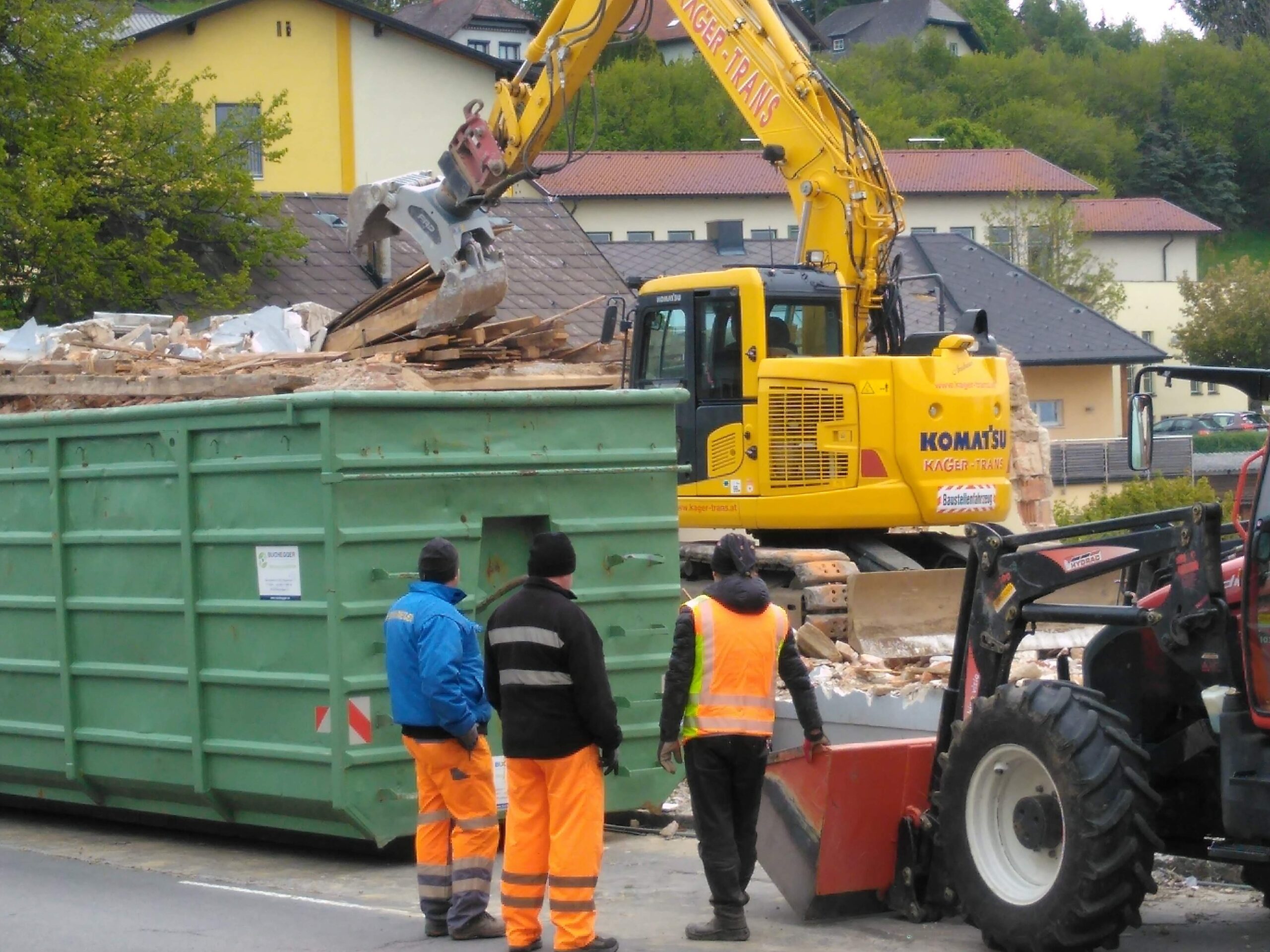 Komatsu-Bagger lädt Abbruchmaterial in einen grünen Container; Arbeiter und Radlader stehen davor.