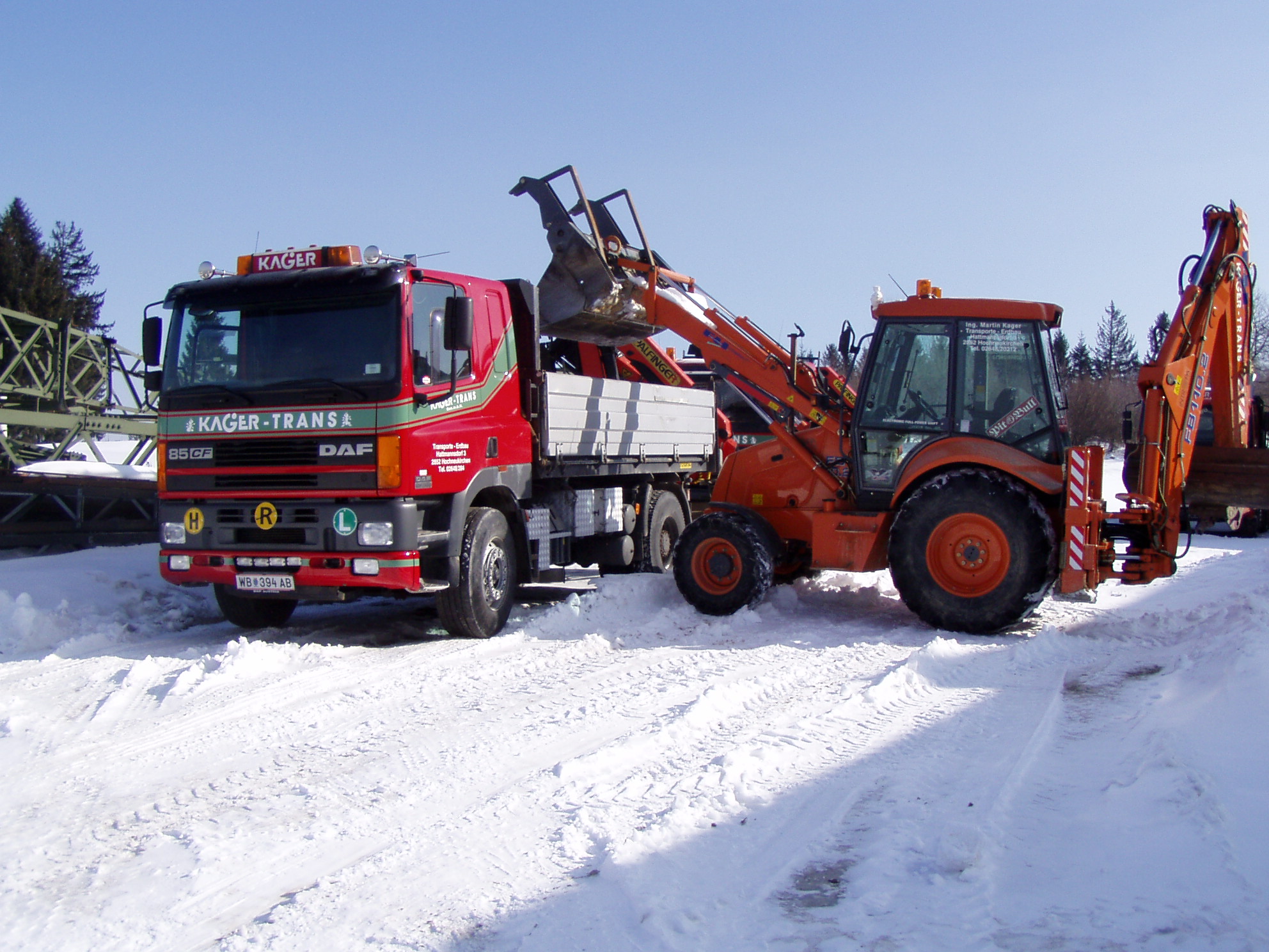 Ein gelber Bagger lädt Schnee auf die Ladefläche eines roten Lkw. 