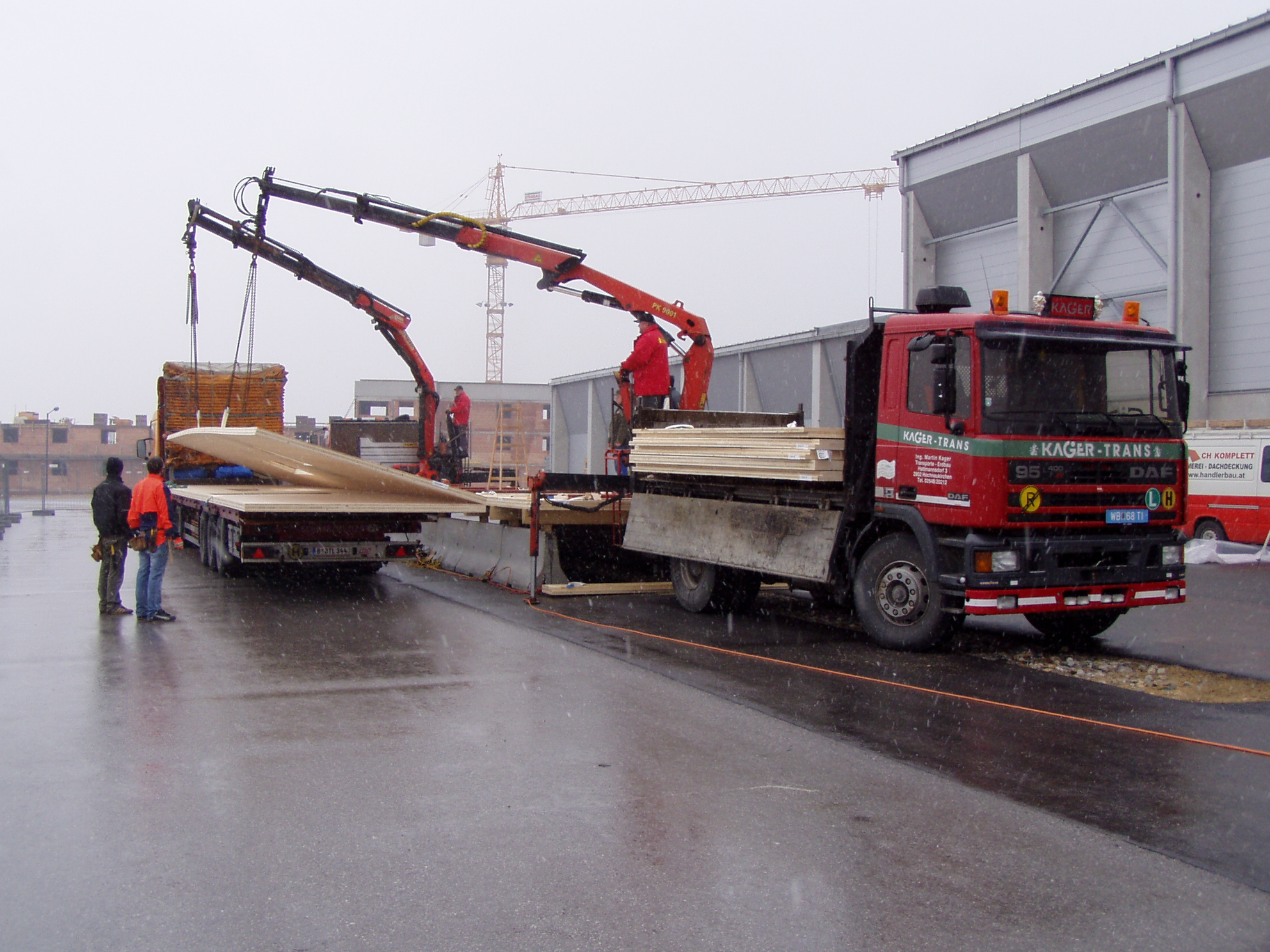 Roter Kager-Trans-Lkw mit zwei Ladekränen entlädt Holz- und Bauelemente auf einer Baustelle; Arbeiter stehen daneben, nasse Fahrbahn und Industriebauten im Hintergrund.