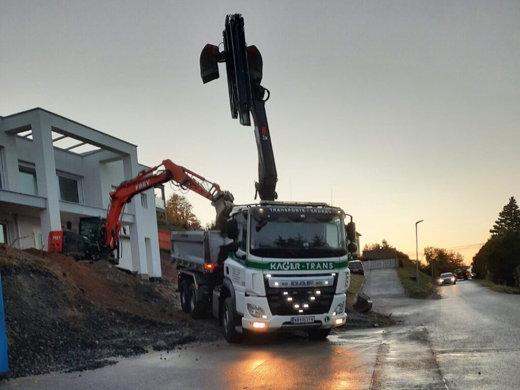 LKW mit Ladekran und Bagger beim Erdaushub vor modernem Wohnhaus bei Sonnenaufgang